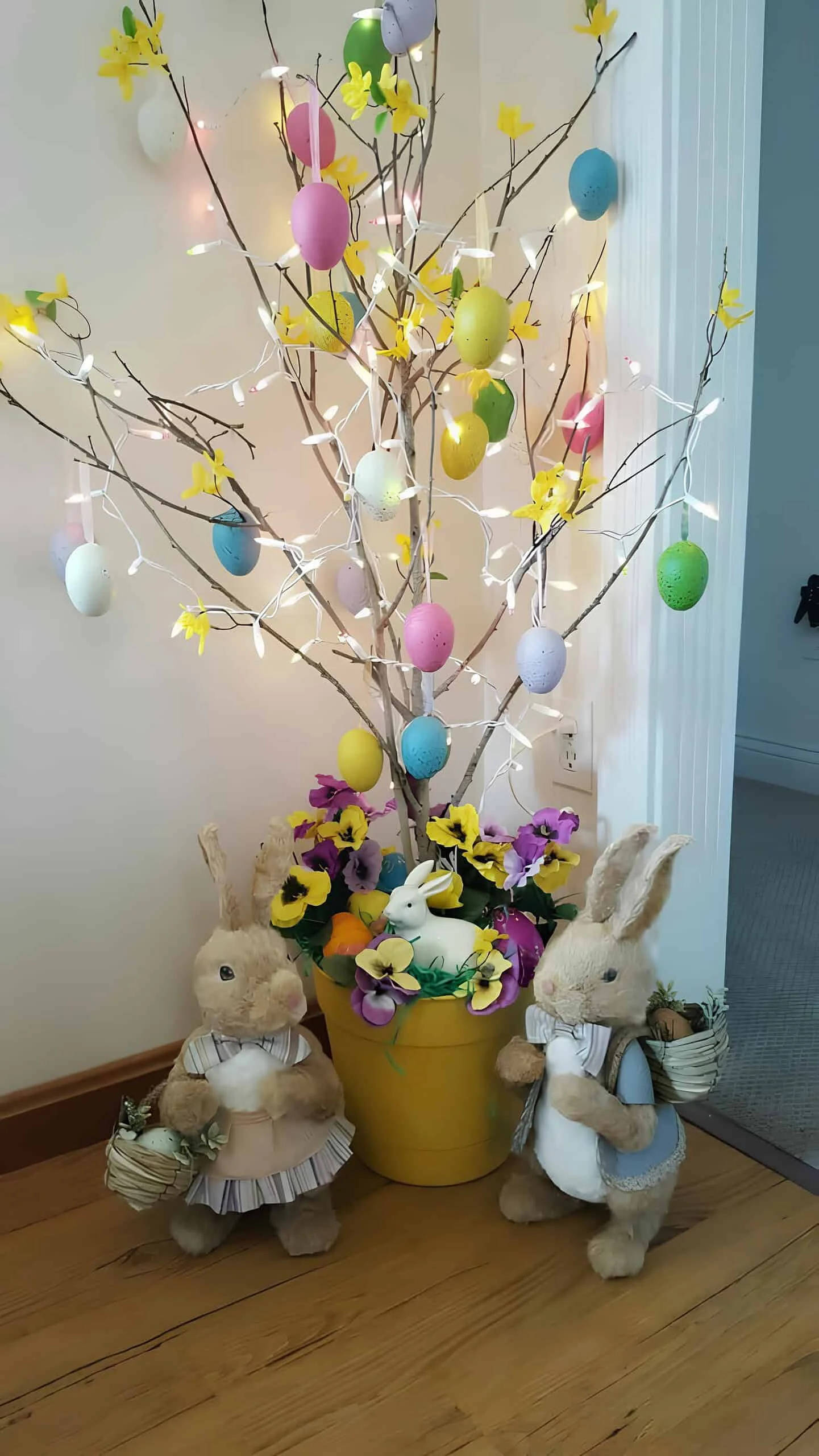DIY árbol de Easter hecho con ramas, huevos colgantes pastel, acentos florales amarillos y luces LED en un macetero amarillo rodeado de conejos de peluche e flores artificiales primaverales - decoración festiva interior para Easter