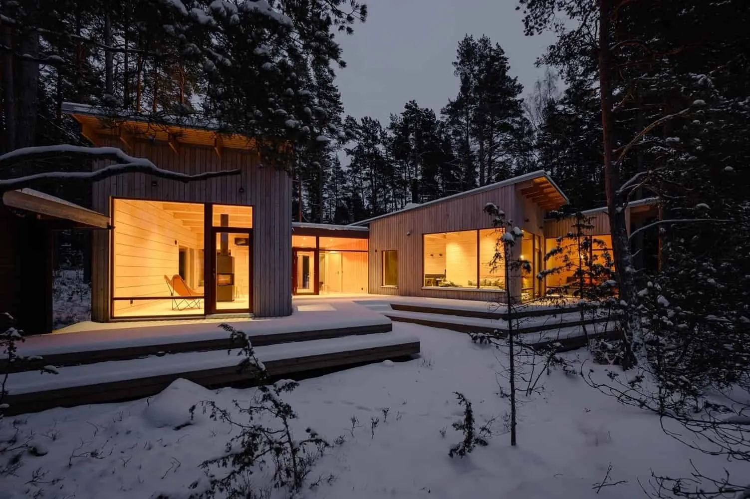 Modern wooden architecture of a cottage in snowy forest surroundings with large glass windows and warm interior lighting, modern design of a mountain estate from Architecture Art Designs.