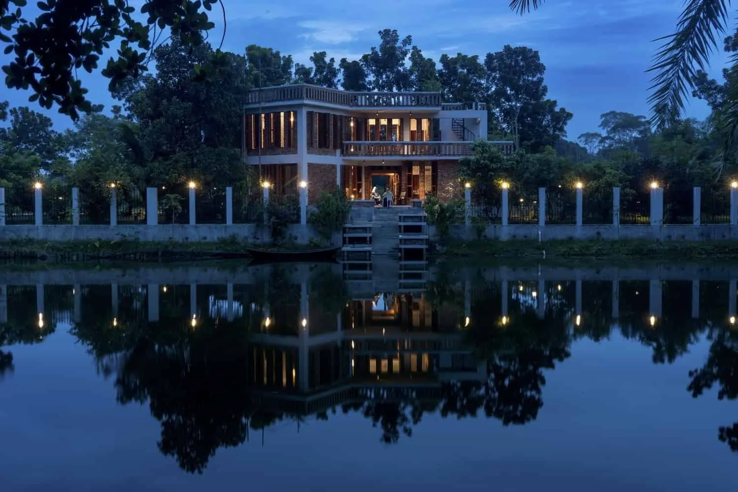 Современный дом у озера с современной архитектурой, окружённый зелеными насаждениями и отражающей водой на закате.