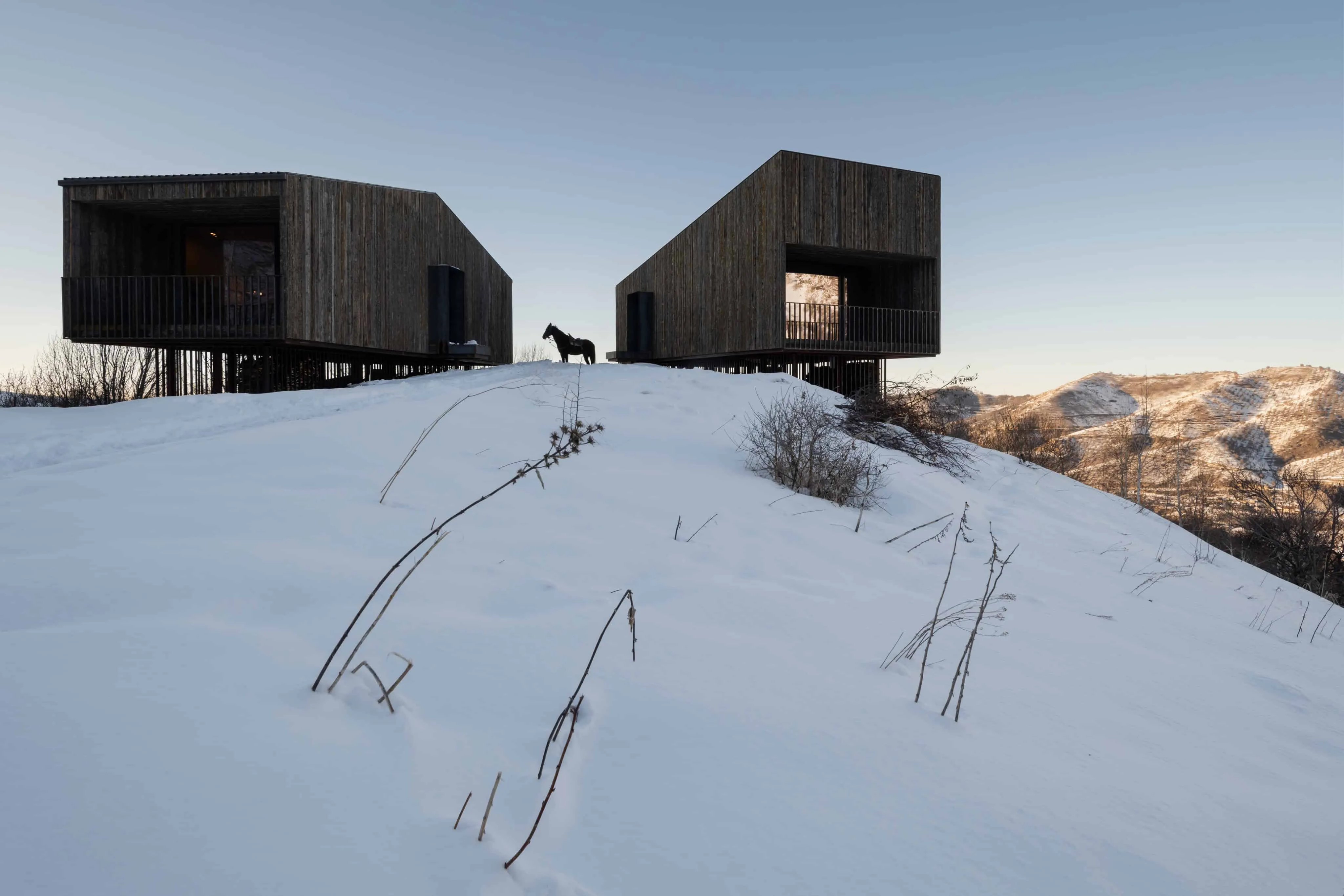 极简木制阿姆小屋矗立在雪地上，旁边有一匹马。