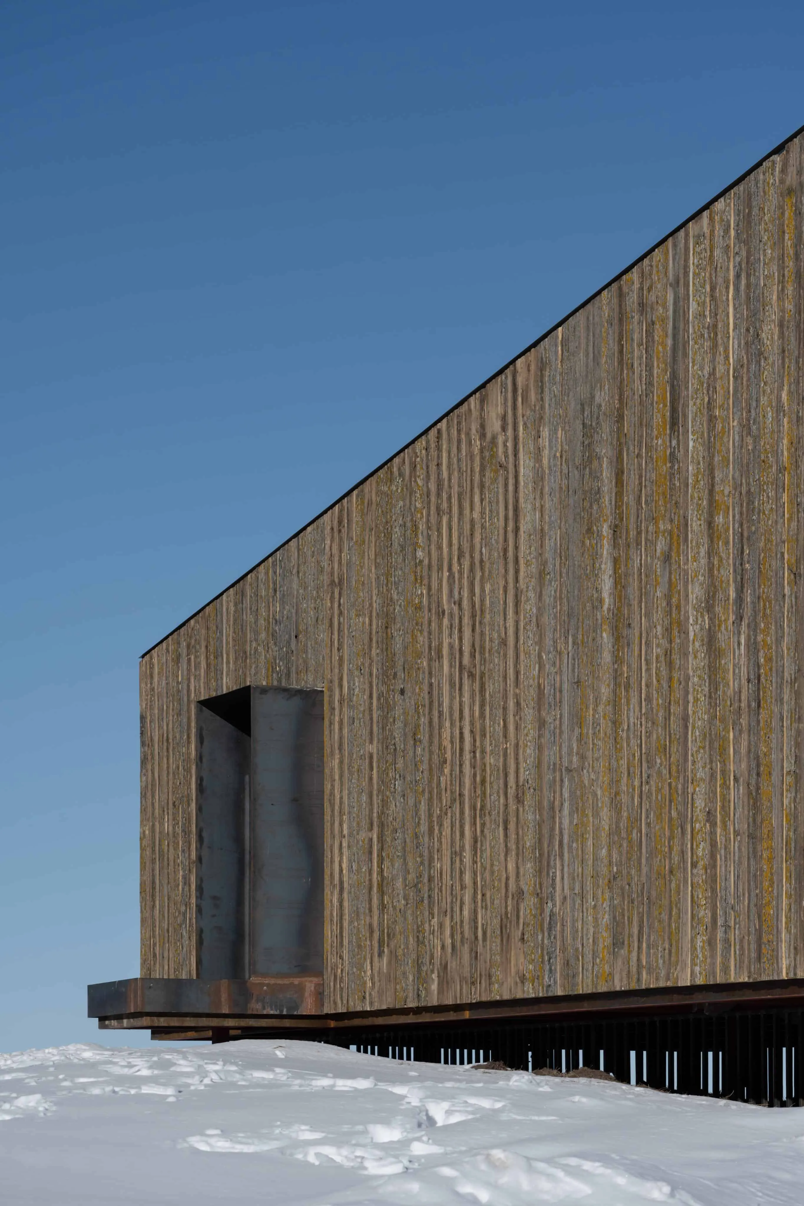 现代阿姆小屋配有垂直木饰面和打开的钢门，在雪景中。