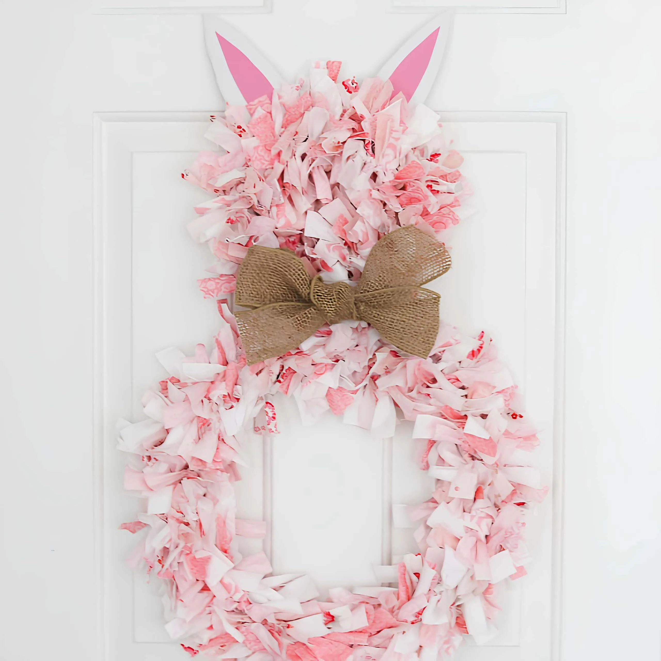 手工制作的粉色布料兔子门饰，用柔软的布条、粗麻丝带和粉色纸耳朵制成，挂在白色门上。