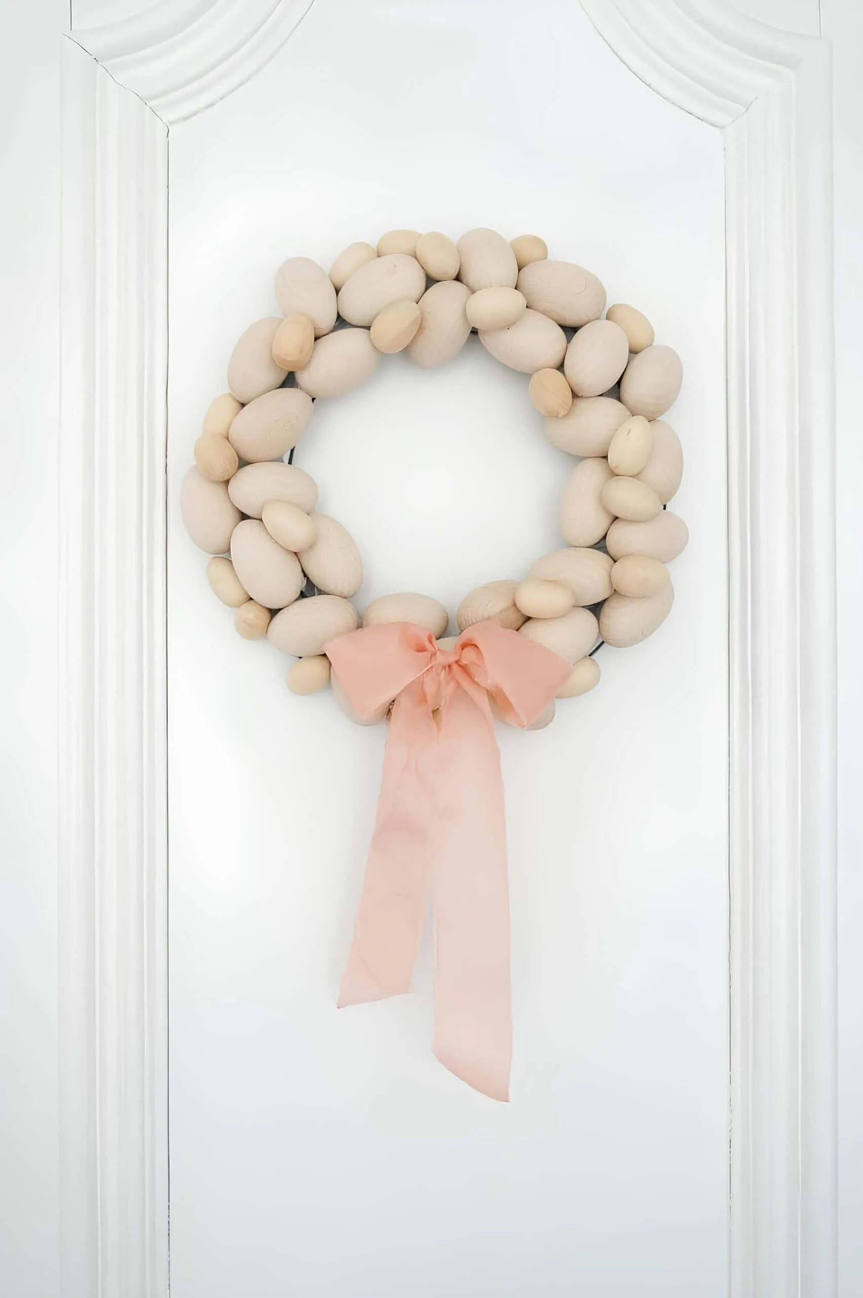 极简鸡蛋门饰，由天然木制鸡蛋制成，并配有柔软的桃色丝带，悬挂在白色门上。