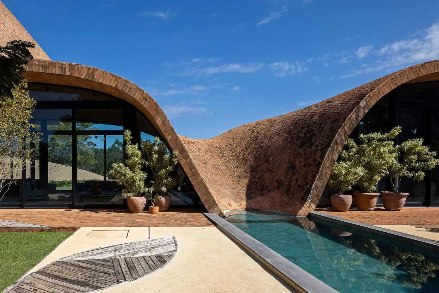 Passive house by Mareines Arquitetura: Self-sufficient cottage in Brazilian rural areas Modern house with unique curved brick architecture and outdoor pool, surrounded by flower pots and abundance of greenery, demonstrating innovative design and stylish outdoor relaxation zones.