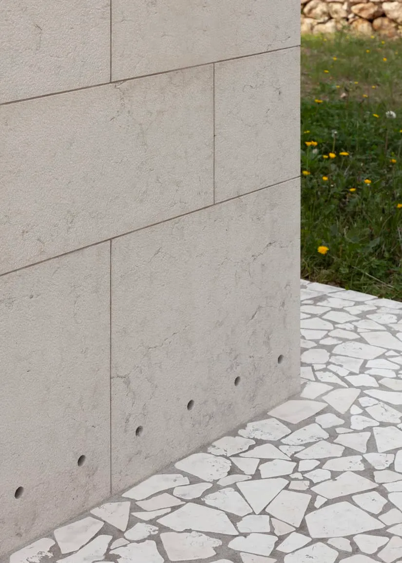 Pared de piedra beige lisa con superficie texturizada y pequeñas grietas, conduciendo a una callejuela de piedra blanca irregular con césped verde denso y pequeñas flores amarillas