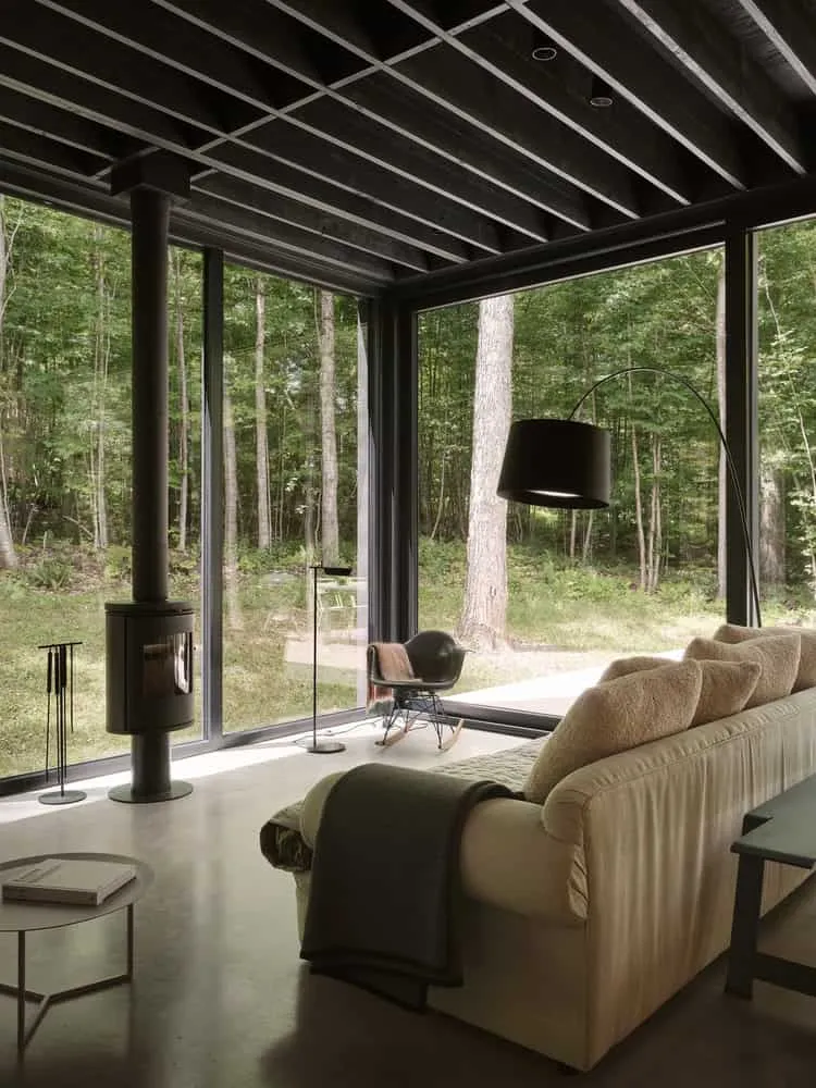 Maison Spiron — angle salon avec cheminée, grandes fenêtres et forêt estivale à Potton, Québec, Canada