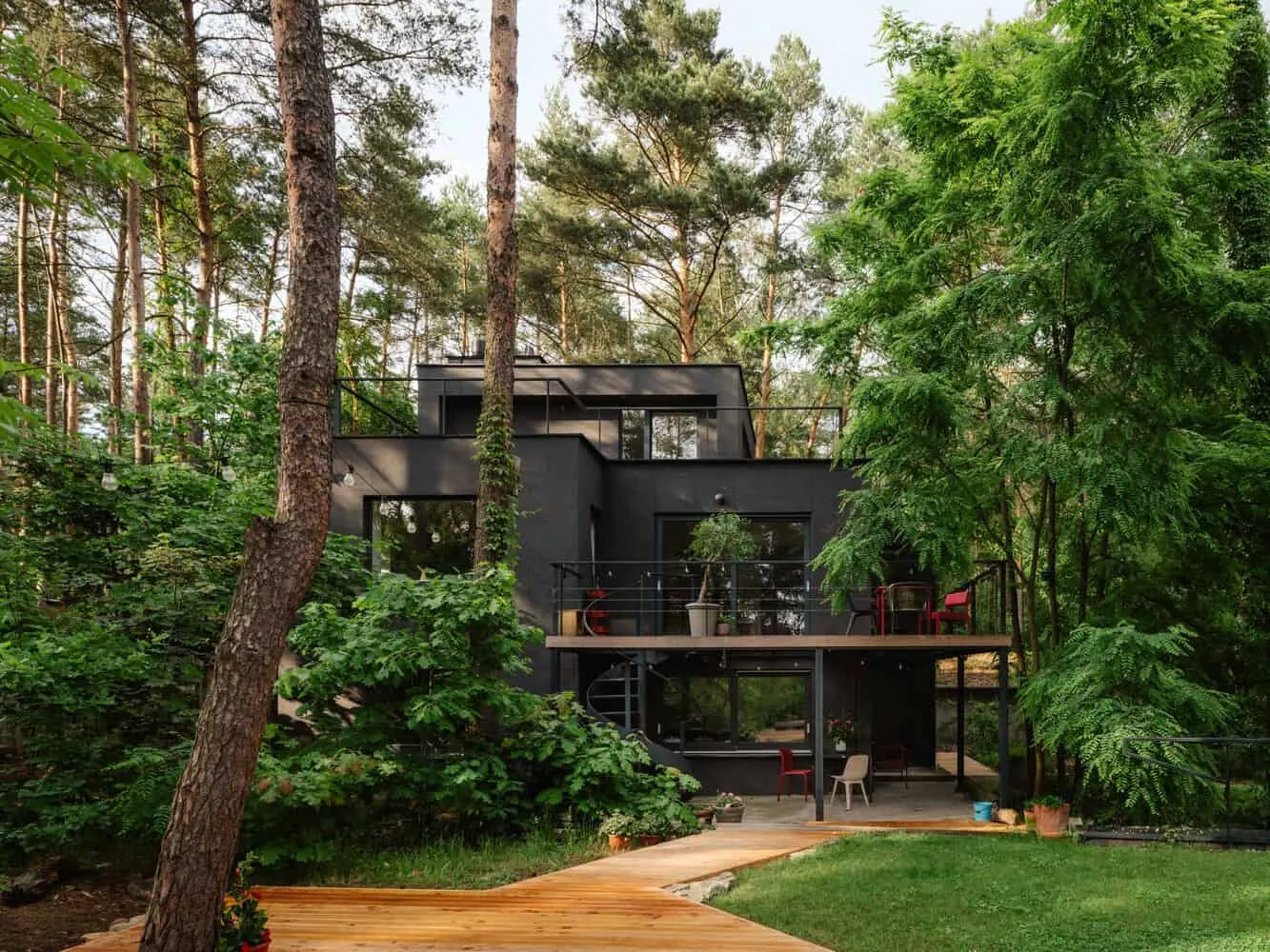 Ein modernes schwarzes Haus mit mehreren Etagen und großen Fenstern, das inmitten hoher Bäume in einem Waldgebiet liegt und über Außenterrassen sowie einen Holzweg verfügt, der zum Garten führt.