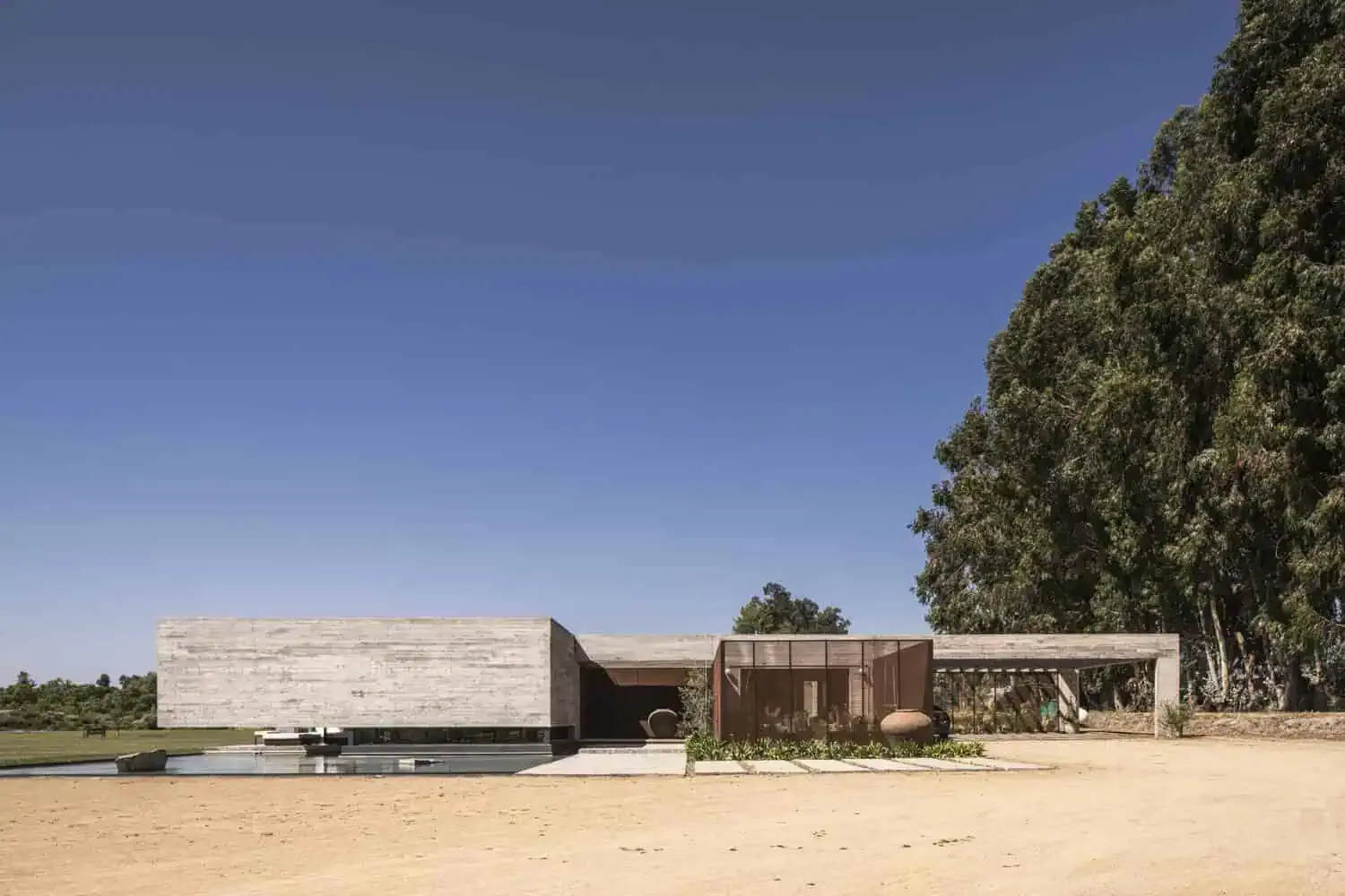 Ein zeitgenössisches minimalistisches Haus mit modernem Architekturdesign, Beton- sowie rostfarbenen Metallakzenten und großen Glasfenstern, gelegen in einer natürlichen Landschaft mit Bäumen und offenen Flächen.