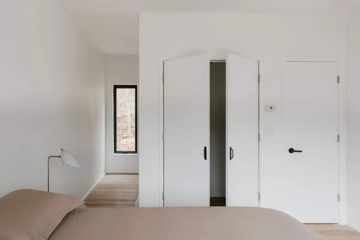 Forest Edge House Bedroom Corridor by Marc Thorpe Design Flur im Schlafzimmer des Forest Edge House mit weißen Schränken und einem schmalen Fenster zum Wald