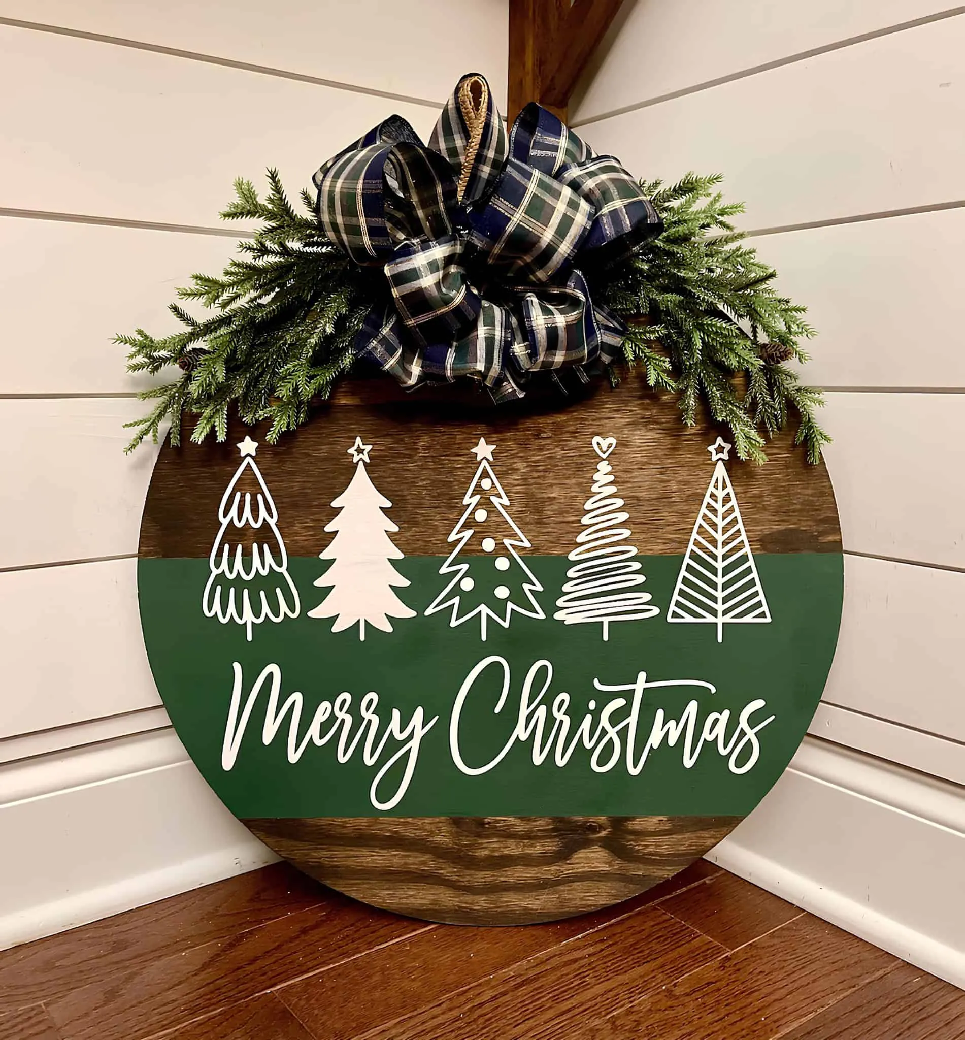 Ländliches rundes Türschild mit kariierter Schleife Rundes Holztürschild mit Grünpflanzen, kariierter Schleife und der Aufschrift ‚Frohe Weihnachten‘