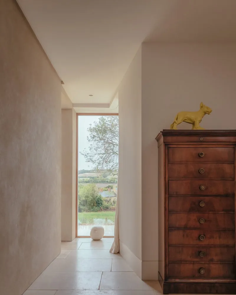 Oliver Leech Architects द्वारा डिज़ाइन किया गया Cotswolds House का न्यूनतमवादी हॉलवे, जिसमें पूरी ऊँचाई वाली खिड़की से प्राकृतिक दृश्य दिखाई दे रहा है