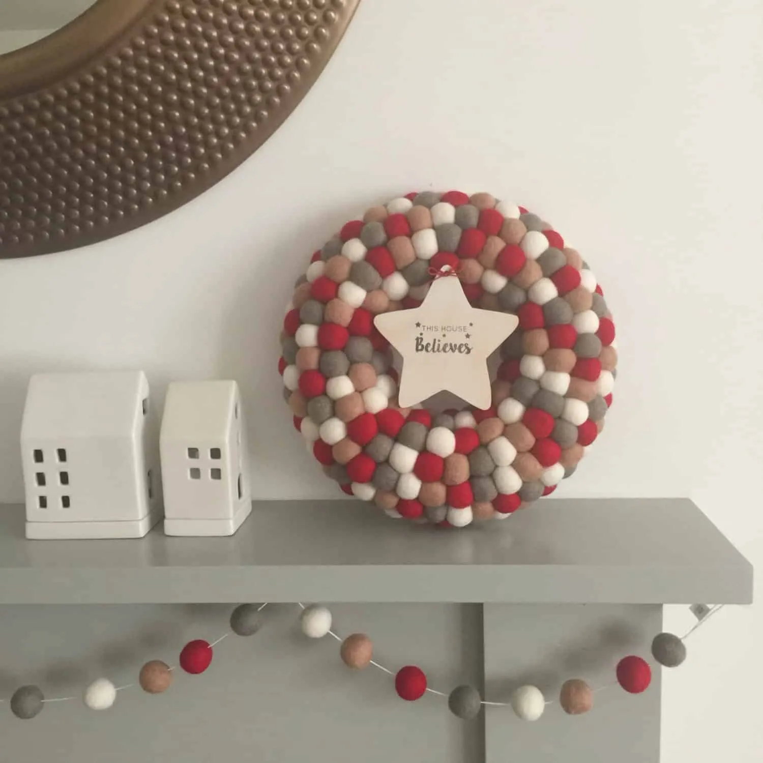 Neutralroter und grauer Pom-Pom-Weihnachtskranz Weihnachtskranz aus neutralroten, grauen und elfenbeinfarbenen Pom Poms mit einem Holzstern-Anhänger auf einer Heizung