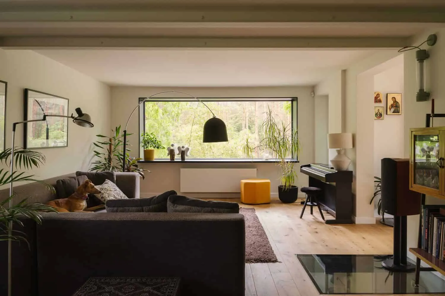 Skubianka House Living Room Panorama by SZCZ Jakub Szczesny Heller, moderner Wohnraum mit großem Fenster, durch das üppige Grünpflanzen zu sehen sind; ausgestattet mit modernen Möbeln, Pflanzen sowie einer Innengestaltung, die natürliches Licht und eine gemütliche Atmosphäre betont.