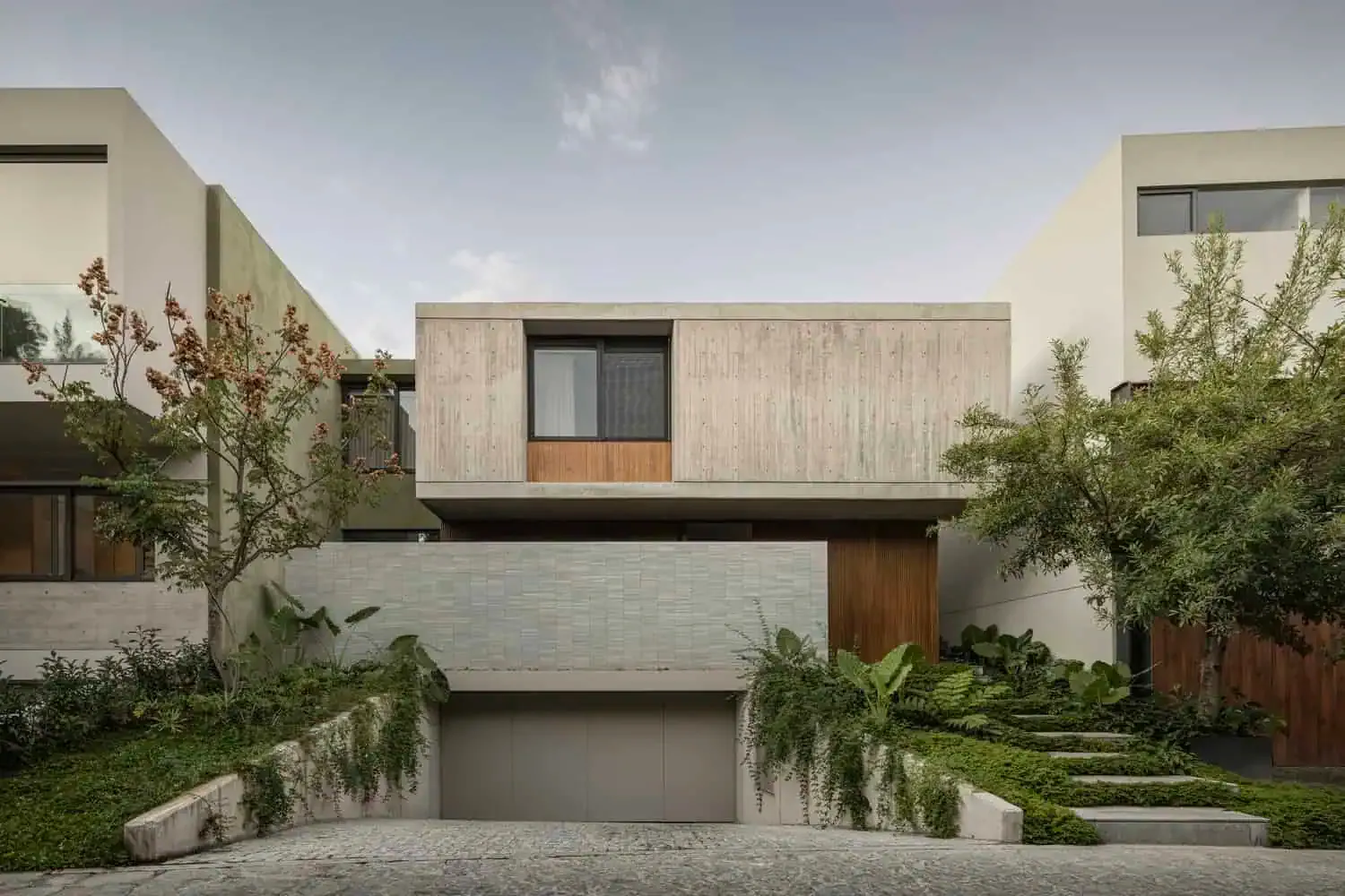 Ein zeitgenössisches, modernes Wohnhaus mit minimalistischer Architektur, Beton- und Holzfassade, üppiger Landschaftsgestaltung sowie großen Fenstern, das innovatives Architekturdesign zeigt.