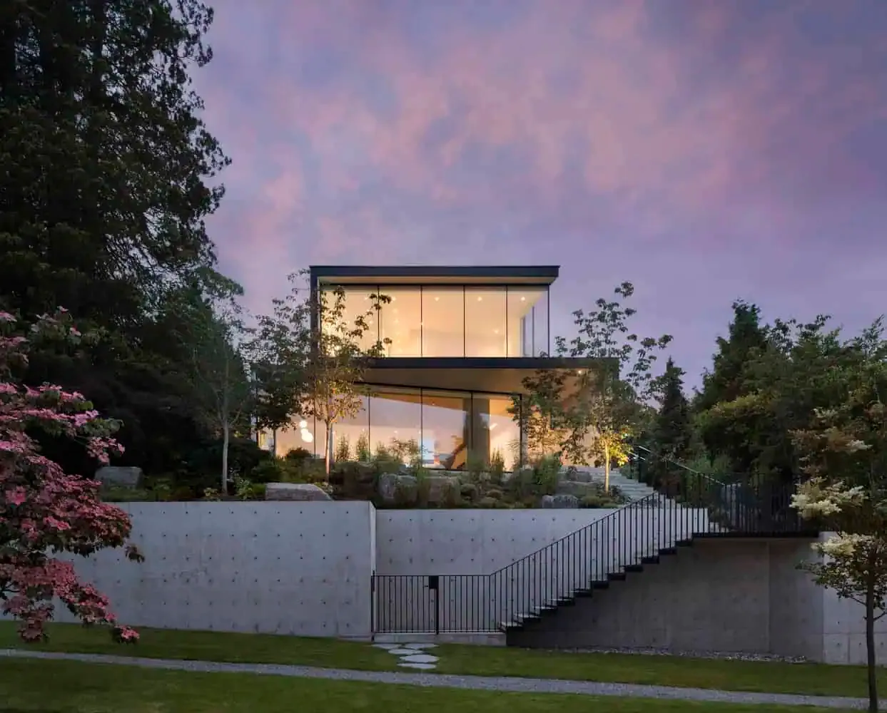 Ein zeitgenössisches modernes Haus mit großen Glasfenstern, umgeben von gestalteten Gärten und Bäumen, mit Blick auf einen farbenfrohen Sonnenuntergangshimmel. Das architektonische Design betont schlanke Linien, offene Räume sowie eine natürliche Integration.