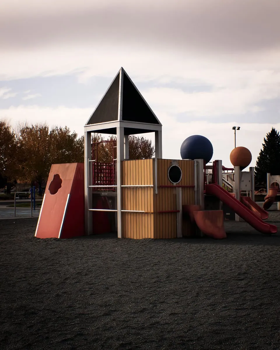 Colorful playground structure with slides, climbing areas, and geometric shapes on a dark textured ground, set against a cloudy sky and autumn trees in a park for children’s outdoor recreation and modern public space design.