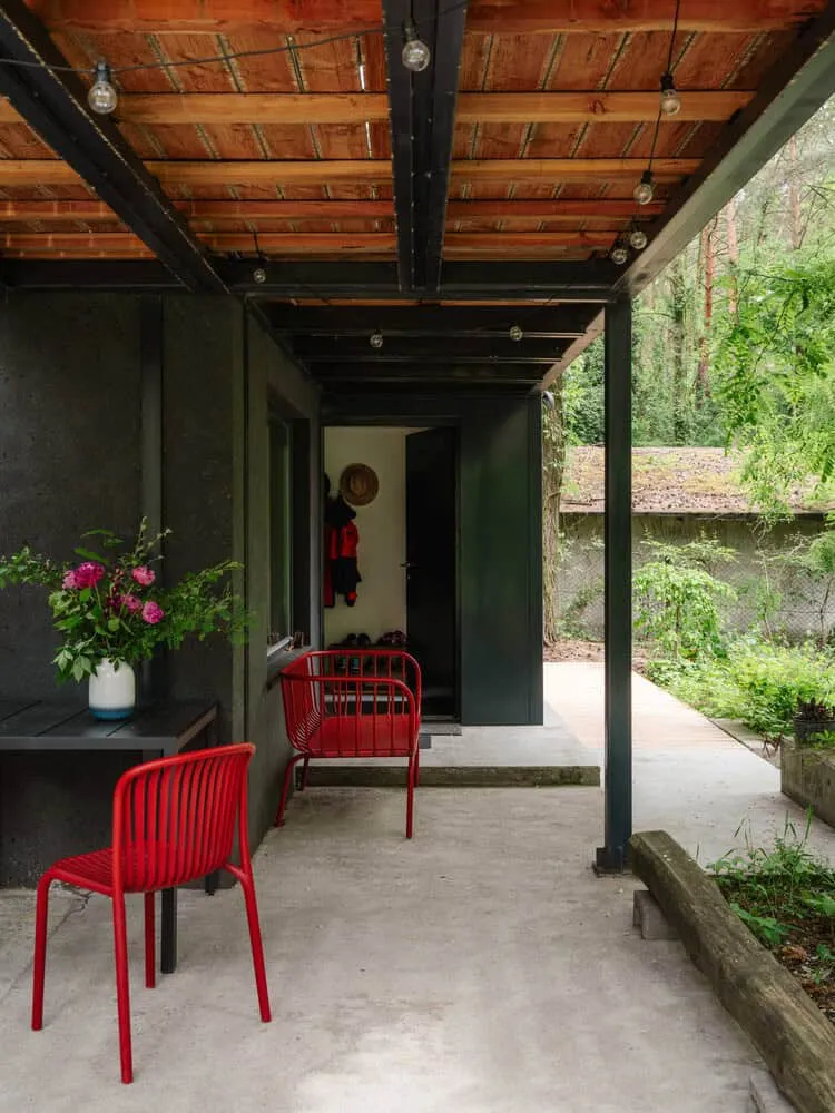 Skubianka House Covered Porch Red Chairs by SZCZ Jakub Szczesny Moderater Außenbereich mit glatten schwarzen Wänden, Holzdecke, stylischen roten Stühlen und üppiger Grünfläche – ein Beispiel für zeitgenössische Architektur und innovatives Design in Außenbereichen.