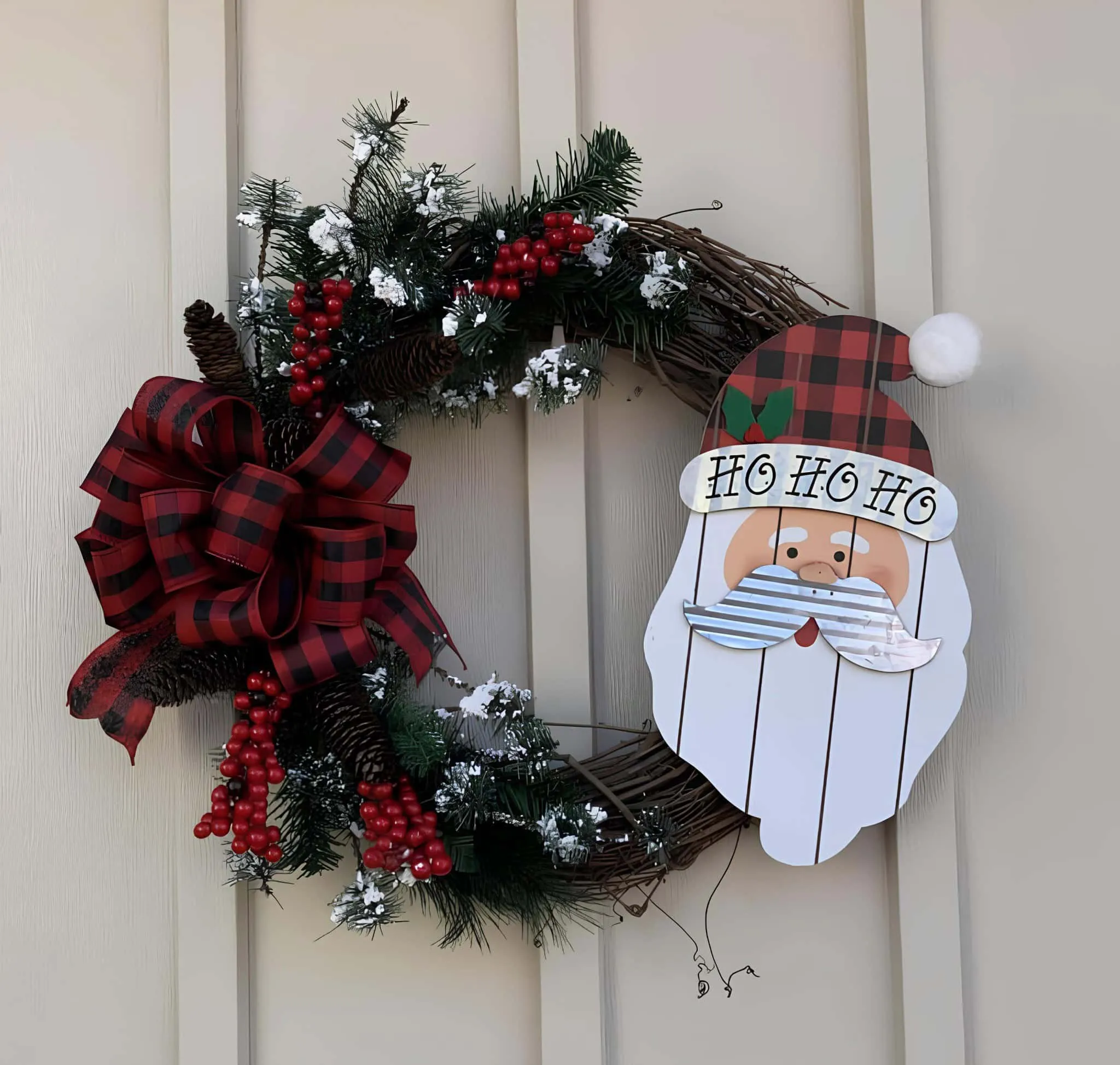Holzkrone zum Weihnachtsfest im Buffalo-Plaid-Stil mit Santa-Motiv Festliche Weihnachtskrone mit Kiefernzweigen, roten Beeren, Kiefernzapfen sowie einem großen roten Plaid-Bogen, die eine Tür zur Feierlichkeiten schmückt.