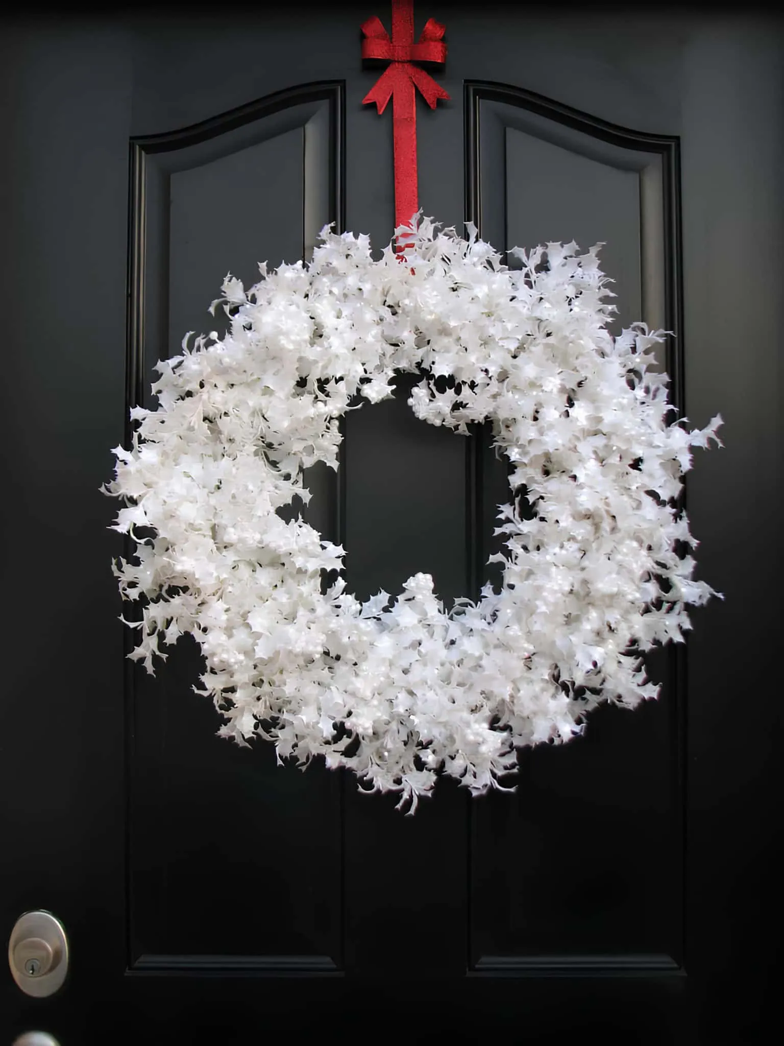 Weißer Feiertagskranz mit rotem Obenbogen Klassische weiße Weihnachtskrone mit einem leuchtend roten Bogen an einer dunklen Tür