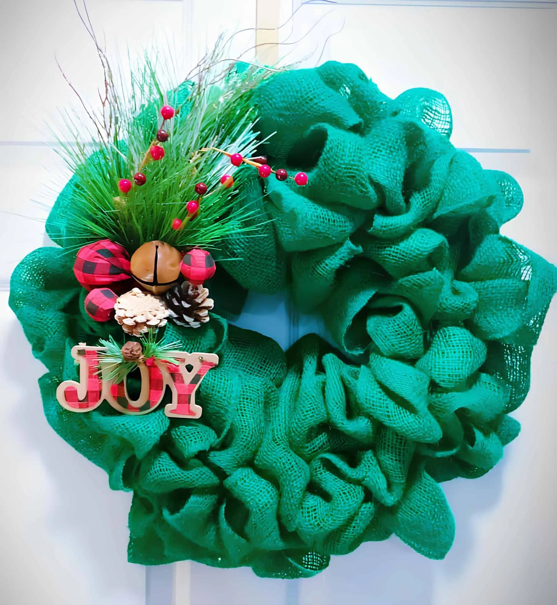 JOY-Schild – Grüner Weihnachtskranz aus Jute Grüner Weihnachtskranz aus Jute mit JOY-Schild, karierten Akzenten, Kiefernzweigen und Glöckchen