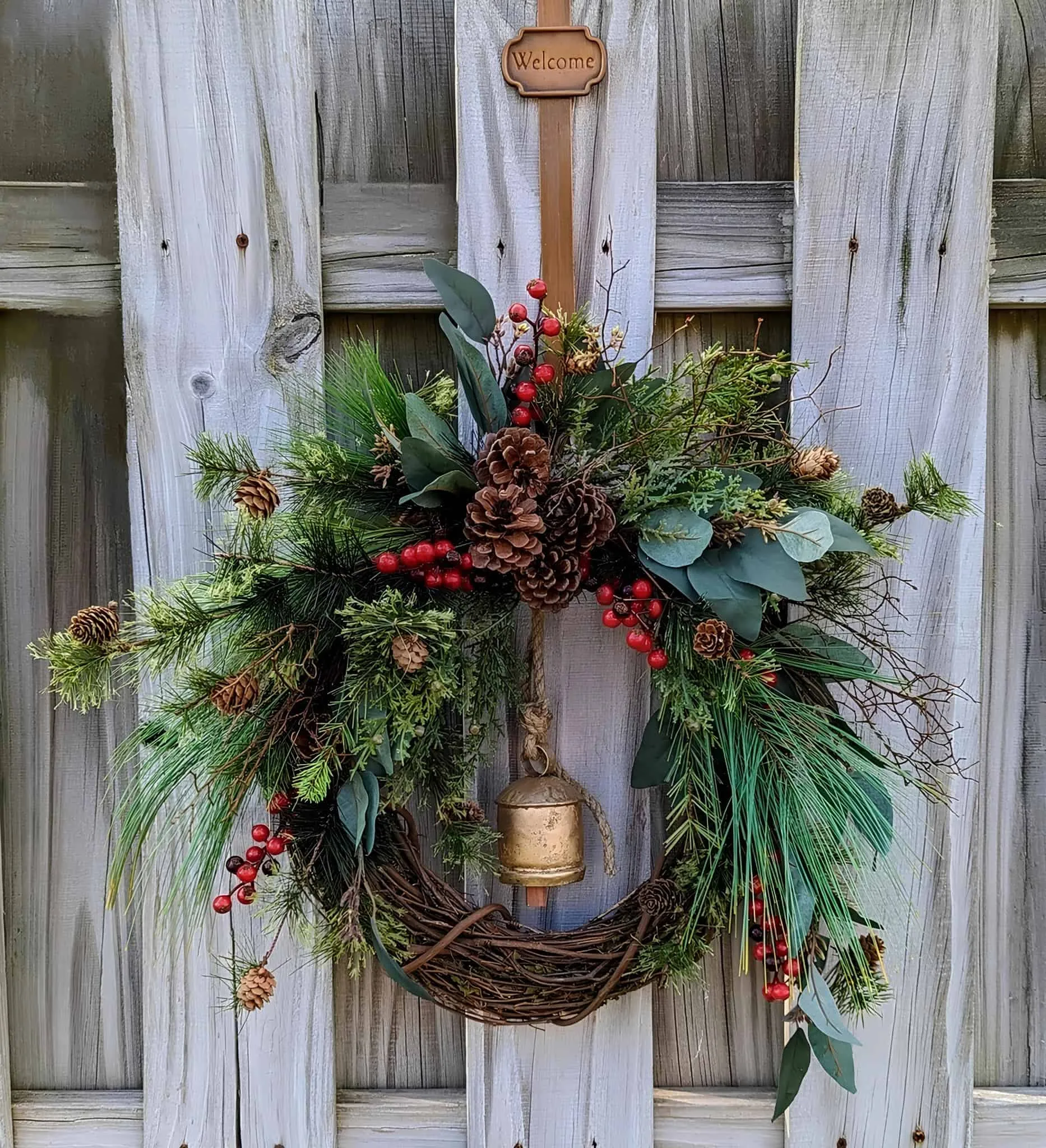 Bell & Pinecone Evergreen Holzweihnachtskranz Lebhafter Feiertagskranz mit Kiefernzapfen, roten Beeren, grünem Laub und einer kleinen Glocke, der an einem verwitterten Holzzaun hängt, um das Zuhause festlich zu dekorieren.