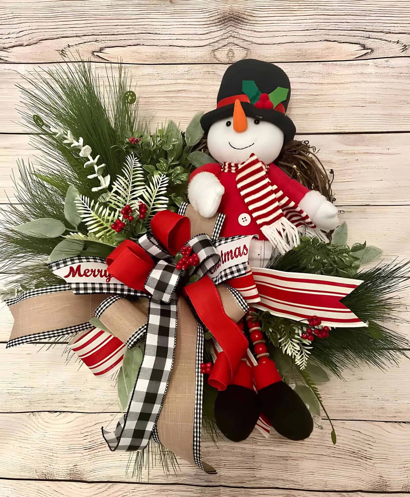 Schneemann-Puppenkranz mit gemischten Bändern Lebendige Weihnachts-Schneemann-Dekoration mit festlichen Bändern, Grünpflanzen und Weihnachtsakzenten auf einem Holzgrund.