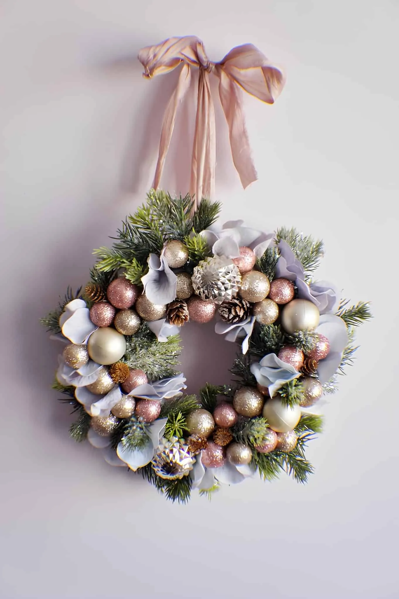 Blush & Gold Ribbon Christmas Wreath Elegante Weihnachtsgirlande, verziert mit pastellfarbenen Ornamenten, Kiefernzapfen und Weihnachtsblättern, die an einem rosa Band hängt und zur saisonalen Wohnraumgestaltung sowie festlichen Innenarchitektur dient.