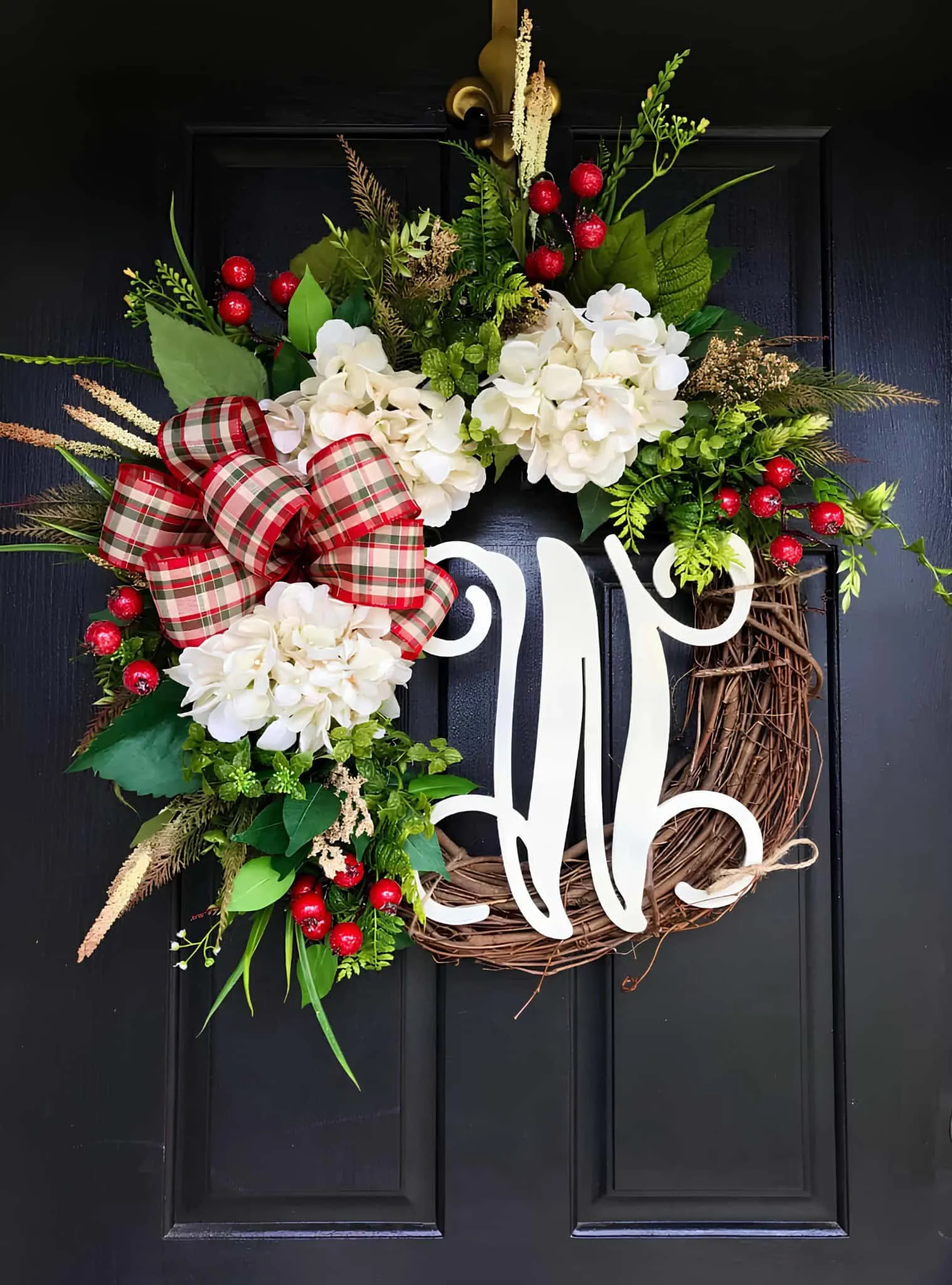 Hydrangea- und Kariertes-Band-Monogramm-Wintergruß aus Holz Lebhafter Weihnachtsgruß mit weißen Hydrangien, roten Beeren, Grünpflanzen und einem karierten Band als Schleife an einer schwarzen Tür, geschmückt für die Feiertage.
