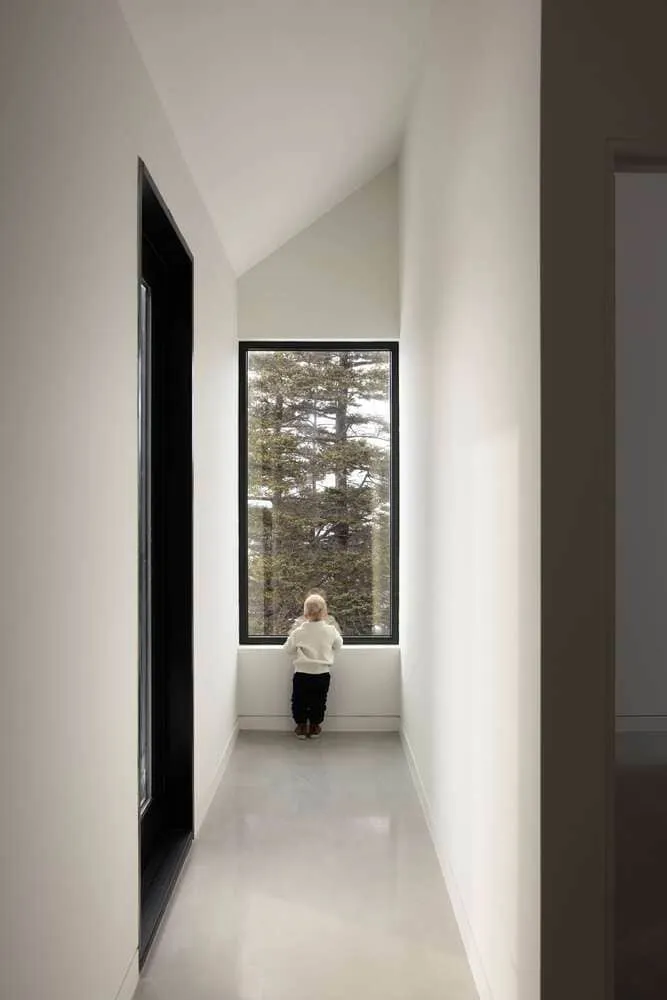 Peter Braithwaite Studio द्वारा डिज़ाइन किया गया Tide Bound House का न्यूनतमवादी आंतरिक गलियारा, जहाँ एक बच्चा ऊँची, जंगल के दृश्य वाली खिड़की से बाहर देख रहा है